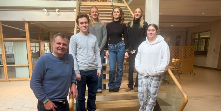 Sex personer står och sitter i en ljus inomhusmiljö vid en trappa, ansikten är suddade.