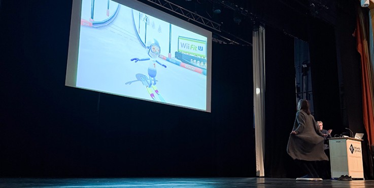 Person spelar Wii Fit U på scen inför publik, med spelet projicerat på stor skärm.