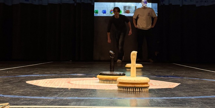 Två personer står på en scen med tre borstar placerade i en cirkel på golvet, bakgrund med mörka gardiner.