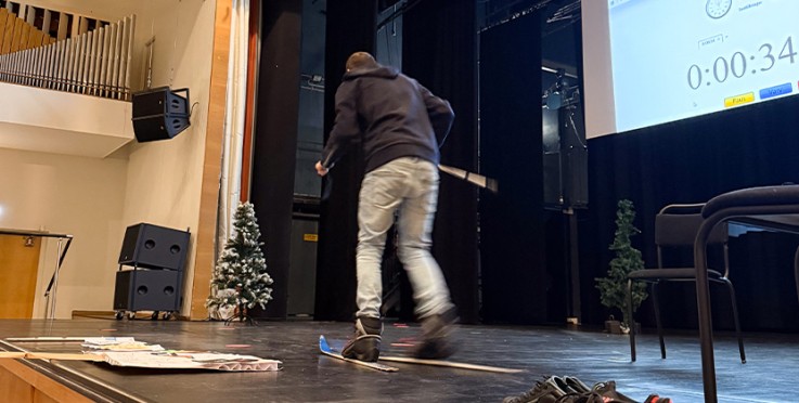 Person på scen med skidor, tidtagarur på skärm, julgranar och stolar i bakgrunden.