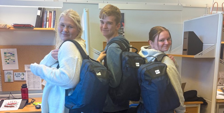 Tre personer med ryggsäckar står i ett kontorsrum med skrivbord och kontorsmaterial.