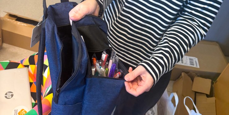Leende person i randig tröja visar upp en blå ryggsäck fylld med pennor i ett kontorsrum.