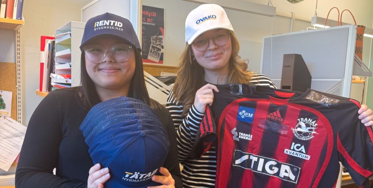 Två personer står i ett kontor, en håller flera kepsar och den andra håller en röd-svart randig sporttröja.