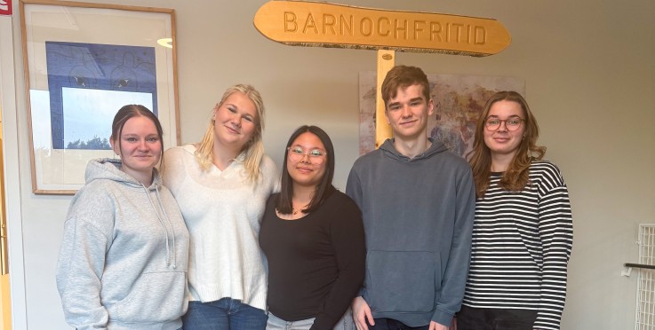 Fem personer står tillsammans inomhus framför en skylt med texten 'Barn och fritid'.