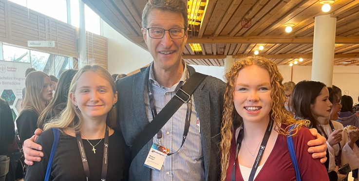 Tre personer poserar tillsammans på en konferens med namnskyltar runt halsen, inomhusmiljö med träpanel i taket.