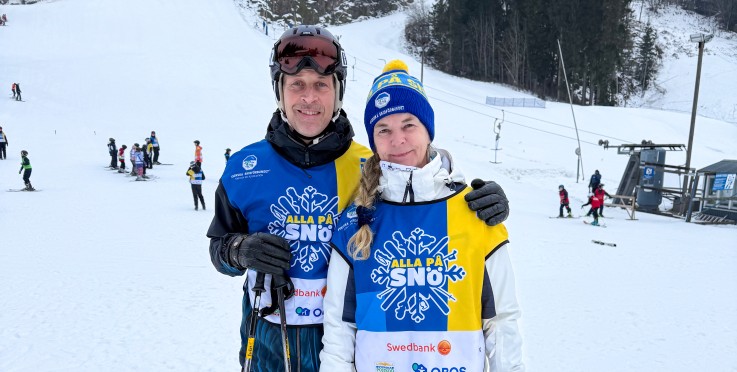 Två personer står på skidor i en snöig skidbacke, omgivna av andra skidåkare och träd.