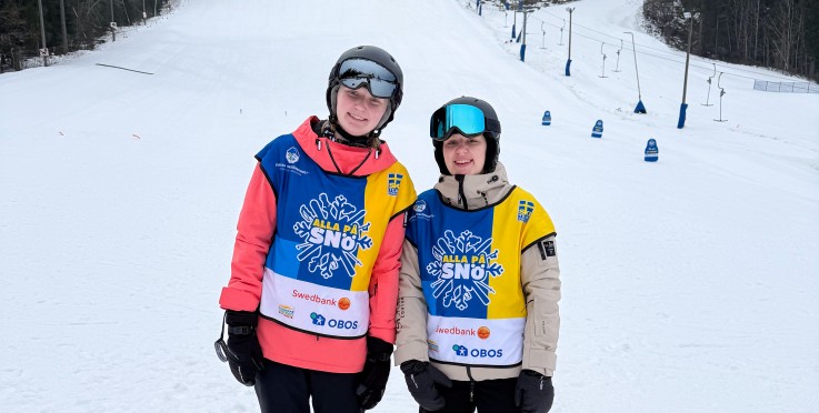 Två personer står på en snöig skidbacke med hjälmar och färgglada västar, omgivna av träd.