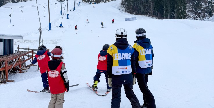 Skidlärare och barn står på en snöig skidbacke, redo att åka skidor. Skidåkare syns längre upp i backen.