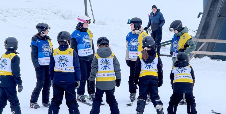 En grupp barn och vuxna står på en snöig skidbacke, klädda i hjälmar och färgglada västar.