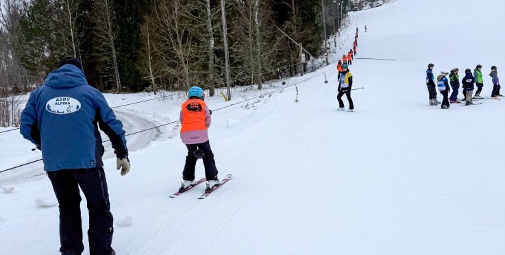 Skidlärare och barn på en snöig skidbacke, flera barn står i kö för att åka lift.