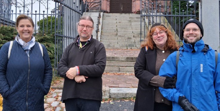 Fyra personer står framför en kyrka med trappor och järnstaket, höstlöv på marken.