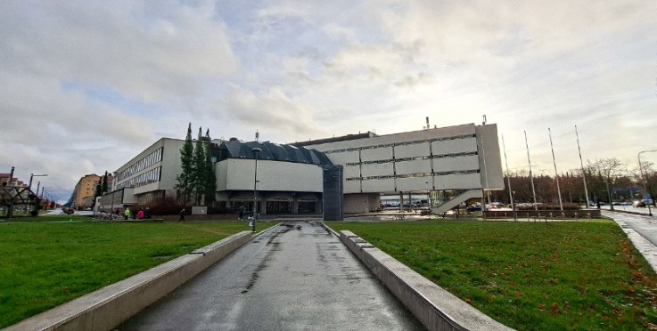 Modern byggnad med grå fasad, omgiven av gräsmatta och gångväg under molnig himmel.