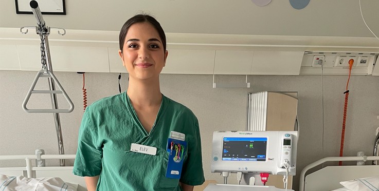 En person i gröna sjukhuskläder står i ett patientrum bredvid medicinsk utrustning.