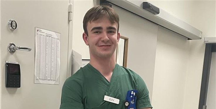 En person i gröna sjukhuskläder står lutad mot en bänk i ett kliniskt rum med armarna i kors.