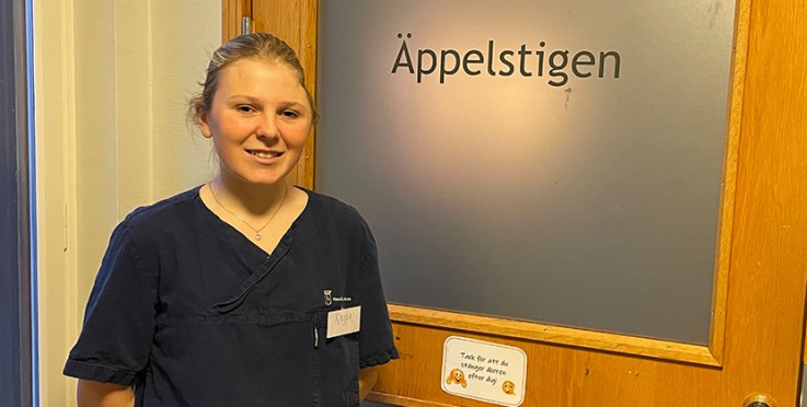 Person i mörk arbetskläder står framför en dörr med skylten 'Äppelstigen'.