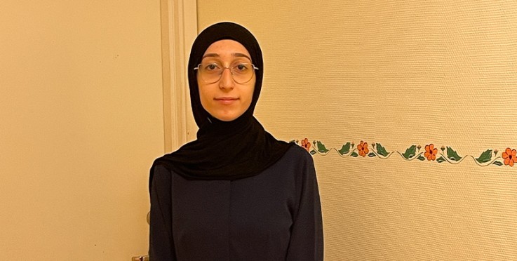 Person med svart hijab och mörk klänning står framför en ljus vägg med blommig bård.