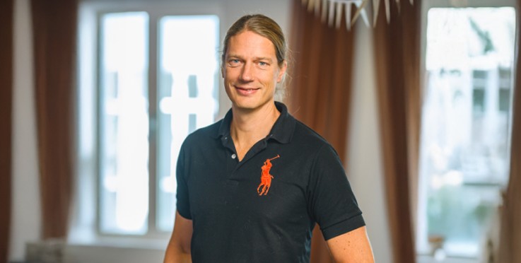 Person med svart pikétröja står i ett rum med stora fönster och bruna gardiner. Ansiktet är suddat.