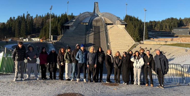 En grupp människor står framför en stor skidbacke utomhus, omgivna av skog och blå himmel.