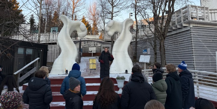 En grupp människor står utomhus framför två vita abstrakta skulpturer och lyssnar på en guide.