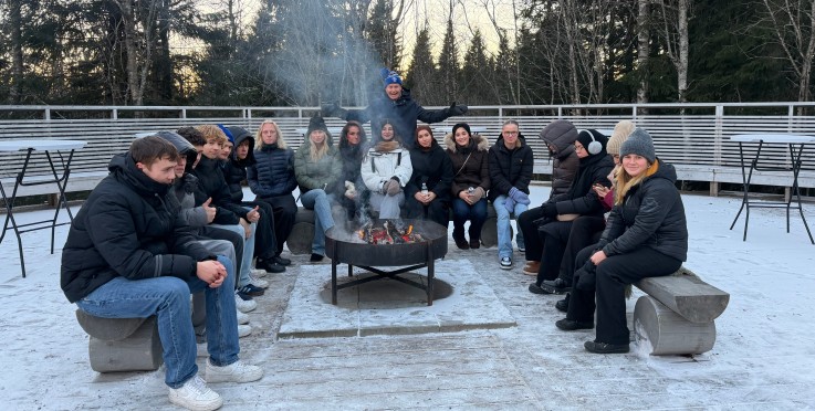 En grupp människor sitter runt en eld utomhus på en snöig plats, omgivna av träd och bänkar.