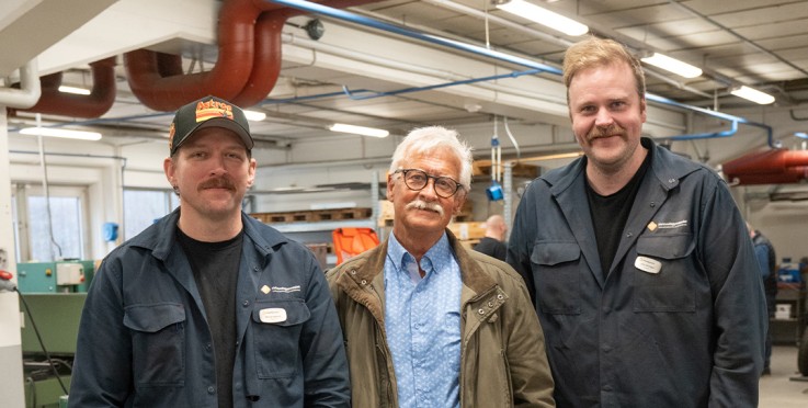 Tre personer står tillsammans i en industrilokal med maskiner i bakgrunden.