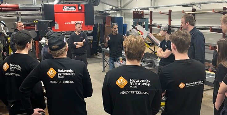 En grupp elever i svarta tröjor står i ett industriklassrum och lyssnar på en instruktör.