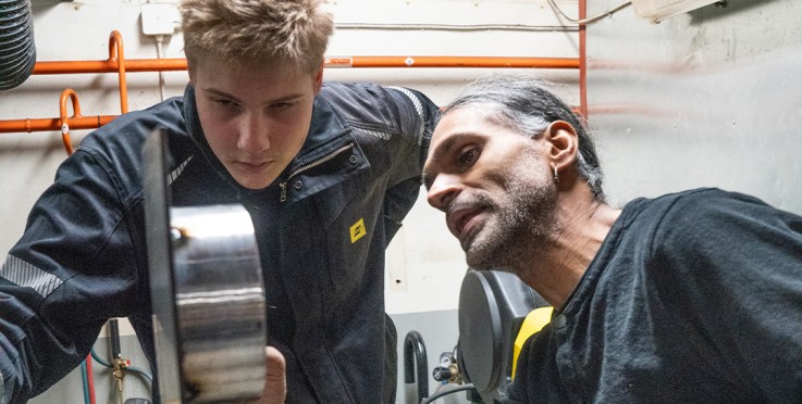 Två personer arbetar tillsammans i ett industriellt verkstadsutrymme med maskiner och metallföremål.