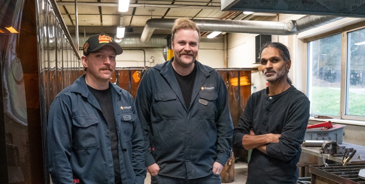 Tre personer står i ett verkstadsrum med arbetskläder, två i blå overall och en i svart tröja.