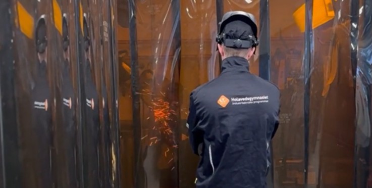 Person i skyddsutrustning svetsar bakom genomskinliga skyddsskärmar, gnistor syns.