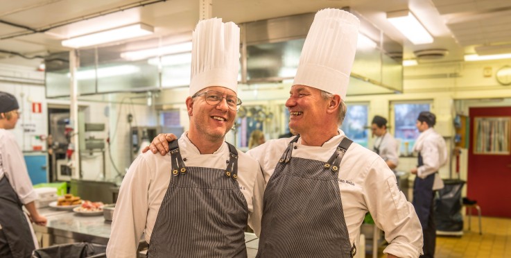 Två kockar med höga vita hattar står tillsammans i ett restaurangkök, omgivna av andra kockar.