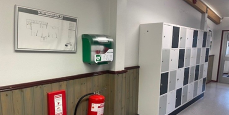 Korridor med brandsläckare, hjärtstartare, utrymningsplan och vita skåp längs väggen.
