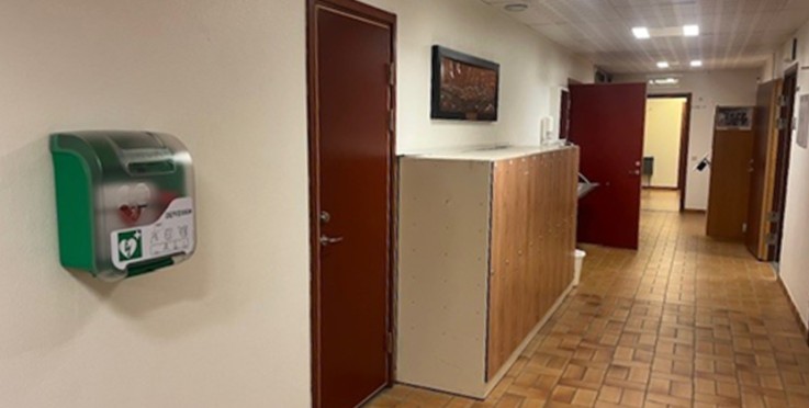 En korridor med bruna dörrar, skåp, hjärtstartare på väggen och klinkergolv.