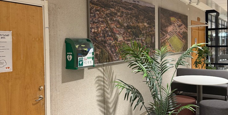 Inomhusmiljö med hjärtstartare på väggen, växter, bord och stolar samt stora flygfoton på väggen.