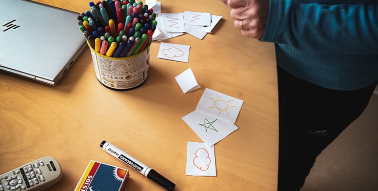 Ett bord med färgpennor, ritade kort, en markerpenna, en fjärrkontroll och en person i blå tröja.
