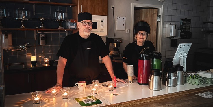 Två personer står bakom en cafédisk med tända ljus, termosar och kaffekoppar.