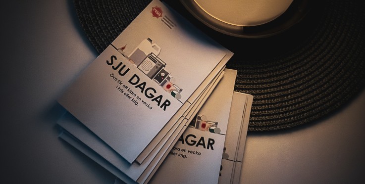 En hög broschyrer med texten 'SJU DAGAR' ligger bredvid ett tänt ljus på ett bord.