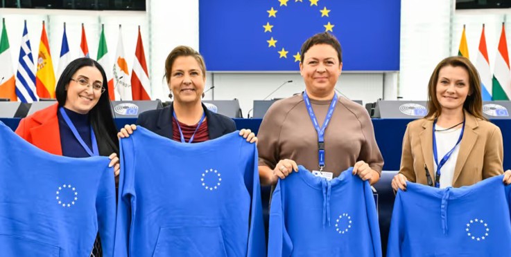 Fyra personer håller blå tröjor med EU-symbolen framför EU-flaggor och podium.