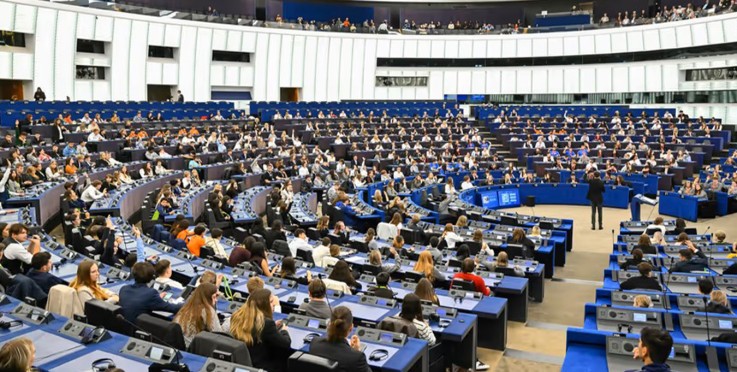 Stor sal med många människor som sitter i rader och lyssnar på en talare, troligen ett parlament.