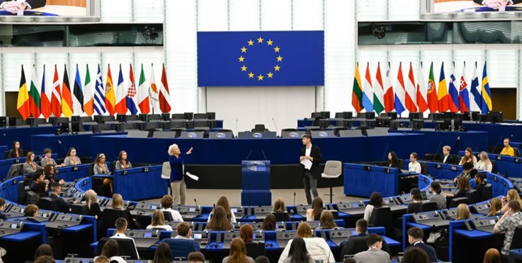 En stor sal med många personer, EU-flaggor och en talare vid podiet, troligen ett möte i Europaparlamentet.