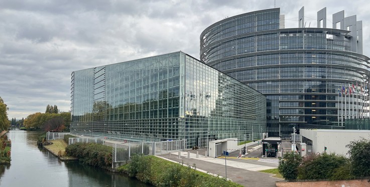 Modern glasbyggnad vid en kanal, med en rund byggnad och flaggor i bakgrunden under molnig himmel.