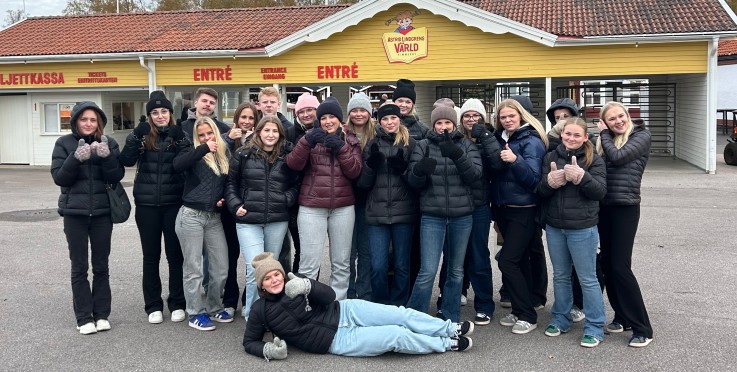 En grupp människor står framför en gul byggnad med skylten 'Astrid Lindgrens Värld'. En person ligger framför gruppen.