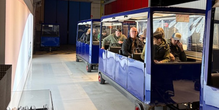 Personer sitter i blå vagn på en inomhus tågattraktion, industriell miljö med takbelysning.