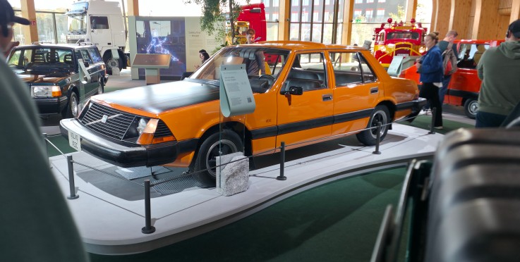 En orange Volvo bil visas på ett museum, omgiven av besökare och andra fordon.