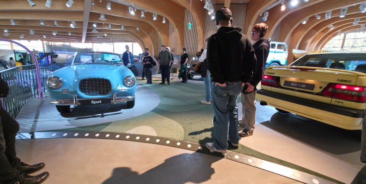 Personer betraktar klassiska bilar, en blå och en gul, i en modern utställningshall med trävalv.