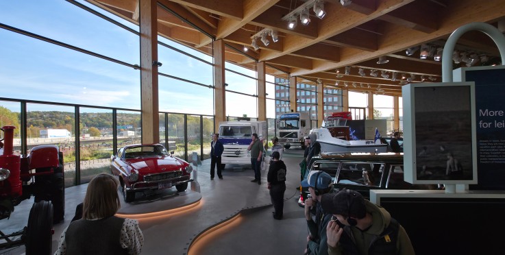 Utställningshall med klassiska bilar, lastbilar och båtar, stora fönster och flera besökare.