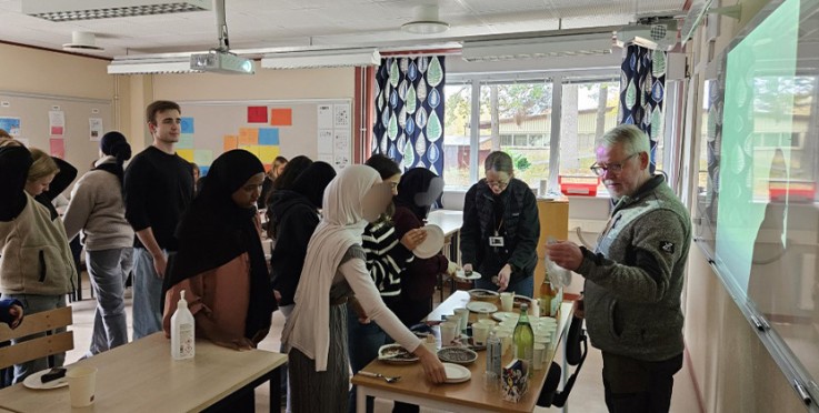En grupp personer samlas runt ett bord i ett klassrum, där mat och dryck serveras.