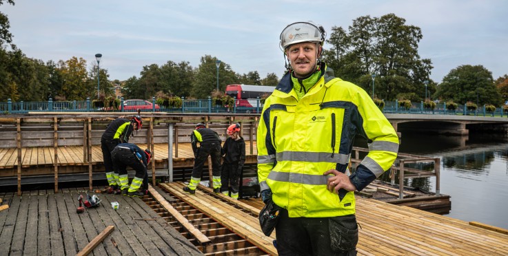 Byggarbetare i reflexkläder arbetar på en träplattform vid vattnet, med bro och träd i bakgrunden.