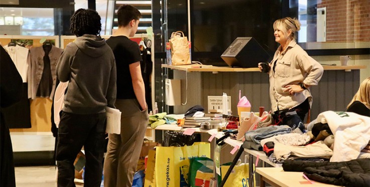 Tre personer står vid ett bord med kläder och prylar, troligen på en loppmarknad inomhus.