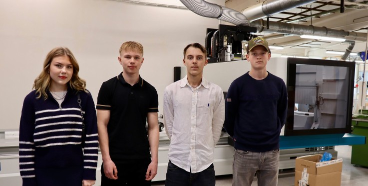 Fyra personer står framför en industriell maskin i en verkstadsmiljö, ansikten är suddade.