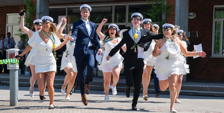 Studenter i vita kläder och mössor springer ut från en byggnad, firar examen.